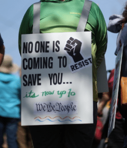 A protest sign worn by a person in a green shirt. The sign reads 'NO ONE IS COMING TO SAVE YOU...' in bold black text at the top, with a raised fist symbol next to the word 'RESIST'. Below, in green cursive writing, it says 'it's now up to' followed by 'We the People' in blue script with a decorative blue wave underneath.